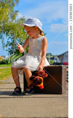 A charming little girl in a white dress and hat is waiting for her vacation. The little girl is holding a plush toy and sitting on an old suitcase, waiting for the vacation train to arrive. The 136965663