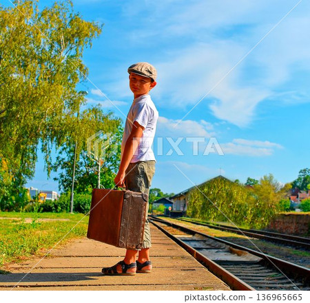A cute little boy holds an old trunk. A cute boy in a baseball cap waits on the station platform for the holiday train. The concept of carefree childhood, travel, adventure, summer vacation, and A cute little boy holds an old trunk. A cute boy in a baseball cap waits on the station platform for the holiday train. The concept of carefree childhood, travel, adventure, summer vacation, and 136965665