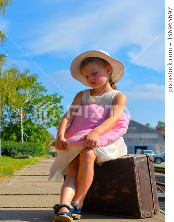 A charming little girl in a white dress and hat is waiting for her vacation. The little girl is holding a plush heart and sitting on an old suitcase, waiting for the vacation train to arrive. The A charming little girl in a white dress and hat is waiting for her vacation. The little girl is holding a plush heart and sitting on an old suitcase, waiting for the vacation train to arrive. The 136965697