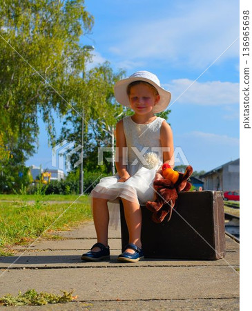 A lovely little girl in a white dress and hat is waiting at the train station for her vacation. The little girl is sitting on an old suitcase, waiting for the vacation train to arrive. The concept of 136965698