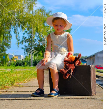 A cute little girl in a white dress and hat waits for her vacation. The little girl sits on an old suitcase, waiting for the vacation train to arrive. The concept of a carefree childhood, summer A cute little girl in a white dress and hat waits for her vacation. The little girl sits on an old suitcase, waiting for the vacation train to arrive. The concept of a carefree childhood, summer 136965711
