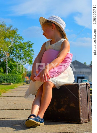 A cute little girl in a white dress and hat is waiting for her vacation. The little girl is waiting for the vacation train to arrive. The concept of a carefree childhood, summer vacation, and family. 136965713
