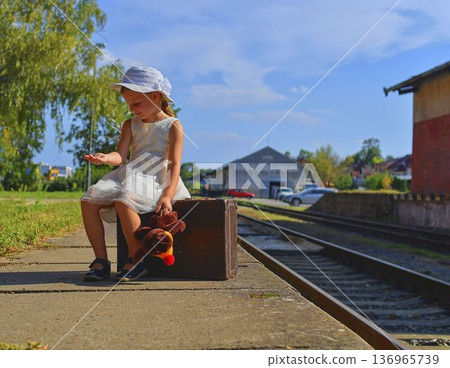 A cute girl at the train station. A little girl holds a plush toy and sits on an old suitcase. The concept of travel, vacations, and a carefree childhood. Retro and vintage vibes. A cute girl at the train station. A little girl holds a plush toy and sits on an old suitcase. The concept of travel, vacations, and a carefree childhood. Retro and vintage vibes. 136965739