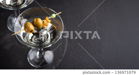 Two glasses with elegant cocktail martini with skewered green olives and a lemon twist on dark textured background Two glasses with elegant cocktail martini with skewered green olives and a lemon twist on dark textured background 136971079