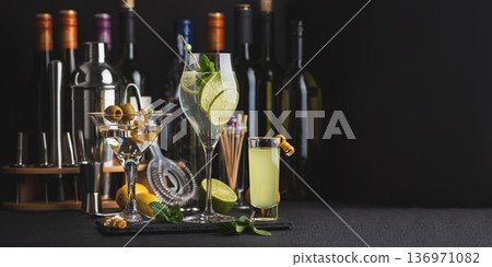 Bar composition featuring a variety of cocktails: martini with olives, citrus mint highball, and limoncello shot against wine bottles, shaker, strainer and fresh ingredients 136971082