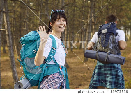 Smiling female hiker with backpack waving while partner walking on forest trail 136973768