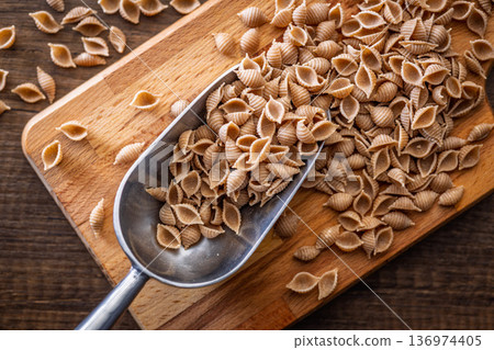 Wholegrain conchiglie pasta on scoop on wooden table. Top view. Wholegrain conchiglie pasta on scoop on wooden table. Top view. 136974405