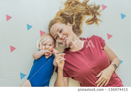 Mother and baby lying on a soft play mat at home, playing, talking and sharing a joyful moment together. Authentic family lifestyle, early childhood development and emotional bonding. Parenting and Mother and baby lying on a soft play mat at home, playing, talking and sharing a joyful moment together. Authentic family lifestyle, early childhood development and emotional bonding. Parenting and 136974415