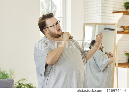 Obese man un glasses and tee singing with comb as microphone at home by mirror 136974444