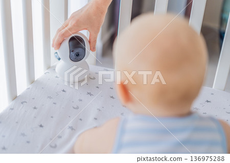 Baby monitor camera placed inside a crib where an infant is lying, ensuring safety and constant supervision. Modern parenting scene combining care and technology. Baby monitoring, security and smart Baby monitor camera placed inside a crib where an infant is lying, ensuring safety and constant supervision. Modern parenting scene combining care and technology. Baby monitoring, security and smart 136975288
