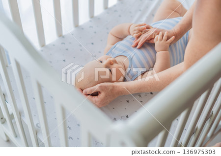 Mother sitting beside the crib gently stroking her baby lying in a cozy bed at home. Warm intimate moment filled with love, trust and calm. Motherhood, infant comfort and emotional bonding concept 136975303