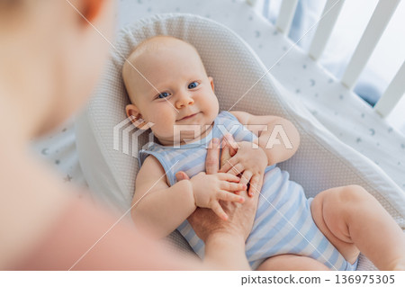 Mother gently sitting beside the crib and stroking her baby resting in a cozy cocoon nest. Tender intimate moment of care and trust at home. Motherhood, infant sleep and emotional bonding concept 136975305