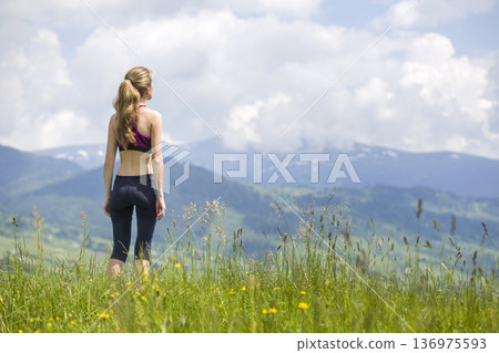 Back view of slim young woman standing on grassy valley on background of green mountains on sunny summer day. 136975593
