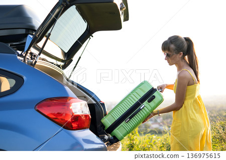 Young female driver putting green suitcase inside her car trunk. Travel and vacations concept. Young female driver putting green suitcase inside her car trunk. Travel and vacations concept. 136975615