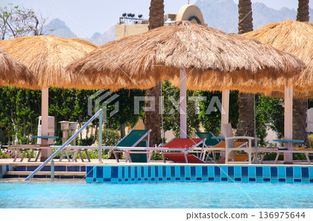 Empty deck chairs under straw shade umbrellas on swimming pool side in tropical resort. Summer vacations and getaway concept 136975644