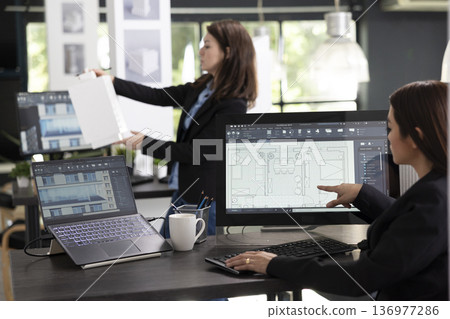 Diverse female professionals working with computer and 3d building model, preparing designs for new contractors. Asian creative designer editing architectural floor plans on touchscreen monitor. Diverse female professionals working with computer and 3d building model, preparing designs for new contractors. Asian creative designer editing architectural floor plans on touchscreen monitor. 136977286