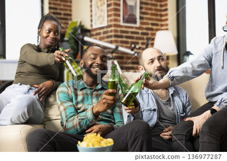 African american and caucasian friends seated on couch, clinking beer bottles and socializing in cozy brick wall apartment. Happy multiethnic group enjoying casual weekend at home. African american and caucasian friends seated on couch, clinking beer bottles and socializing in cozy brick wall apartment. Happy multiethnic group enjoying casual weekend at home. 136977287