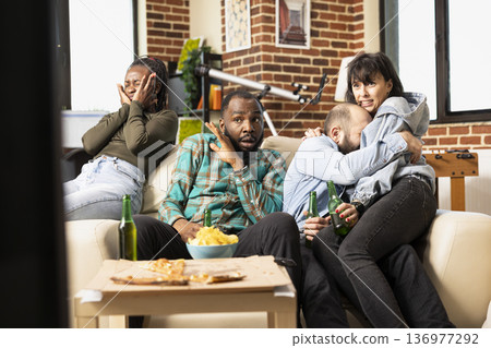 Frightened friends watch horror movie together in apartment, reacting to shocking scene with fear and nervous expressions. Caucasian couple hugs as diverse group bonds over thriller film at home. Frightened friends watch horror movie together in apartment, reacting to shocking scene with fear and nervous expressions. Caucasian couple hugs as diverse group bonds over thriller film at home. 136977292