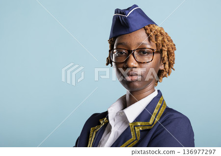 Professional cabin crew member looks at camera, posing confidently in studio. Portrait of elegant black woman in aviation uniform and glasses, representing female confidence and leadership. Professional cabin crew member looks at camera, posing confidently in studio. Portrait of elegant black woman in aviation uniform and glasses, representing female confidence and leadership. 136977297