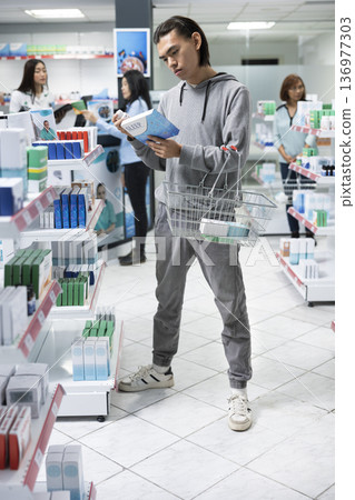 Asian male shopper examines insomnia medications in neighborhood drugstore, reading instructions and ingredients. Young adult carefully pharmaceutical options to support better sleep. Asian male shopper examines insomnia medications in neighborhood drugstore, reading instructions and ingredients. Young adult carefully pharmaceutical options to support better sleep. 136977303