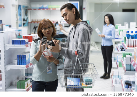 Asian man shows smartphone to pharmacy employee, asking for help finding healthcare product. Female drugstore attendant holds medication and assists male customer based on image on device. 136977340