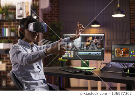 Asian freelancer uses VR headset and editing software to create cinematic montage for film production. Male editor immersed in augmented reality workspace, enhancing video with visual effects at home. 136977362