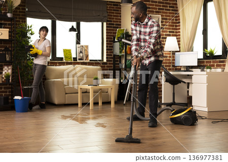 Smiling african american man vacuums hardwood floor while Caucasian wife wipes shelves in modern living room. Diverse couple enjoys teamwork during household chores, maintaining cleanliness at home. Smiling african american man vacuums hardwood floor while Caucasian wife wipes shelves in modern living room. Diverse couple enjoys teamwork during household chores, maintaining cleanliness at home. 136977381