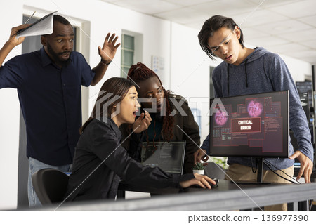 Diverse colleagues show frustration and focus while analyzing system crash on computer screen in tech startup workspace. Agile development team collaborates to solve complex programming bug. 136977390
