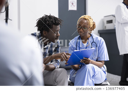 Female physician writes detailed observations while consulting man in hospital reception area. Nurse in scrubs explains health information and reassures patient with focused treatment discussion. Female physician writes detailed observations while consulting man in hospital reception area. Nurse in scrubs explains health information and reassures patient with focused treatment discussion. 136977405