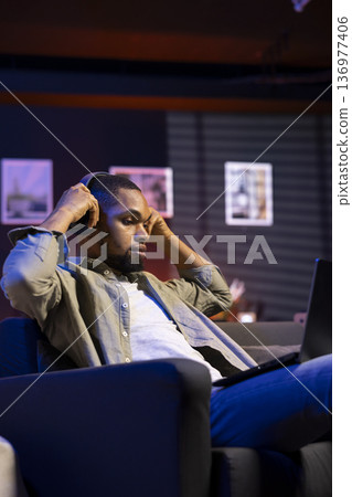 Black self employed guy browsing the web and listening to music, having fun with songs on headset and completing his tasks. African american man laying on sofa and relaxing in the living room. Black self employed guy browsing the web and listening to music, having fun with songs on headset and completing his tasks. African american man laying on sofa and relaxing in the living room. 136977406