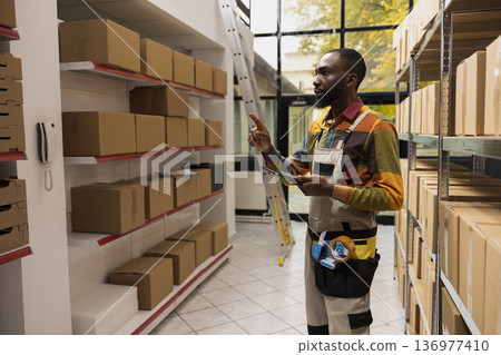 Black man worker counting pallets of goods with inventory list on tablet, product handling and express delivery service for e-business. Retail industry and in house logistics operations. 136977410
