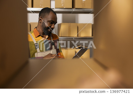 African american depot worker preparing parcels for transportation, handling inventory for e-shop shipments and ensuring product quality control. Support online shopping and express delivery. 136977421