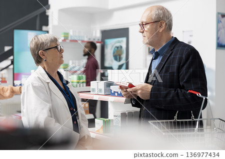 White woman in lab coat assists male customer in local drugstore, providing prescribed medication. Female pharmacist helps senior man by recommending appropriate supplements and healthcare products. White woman in lab coat assists male customer in local drugstore, providing prescribed medication. Female pharmacist helps senior man by recommending appropriate supplements and healthcare products. 136977434