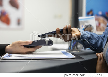 Closeup of contactless payment transaction as african american patient taps phone at hospital front desk. Clinic receptionist holds pos as client uses nfc technology for secure, cashless checkout. Closeup of contactless payment transaction as african american patient taps phone at hospital front desk. Clinic receptionist holds pos as client uses nfc technology for secure, cashless checkout. 136977435