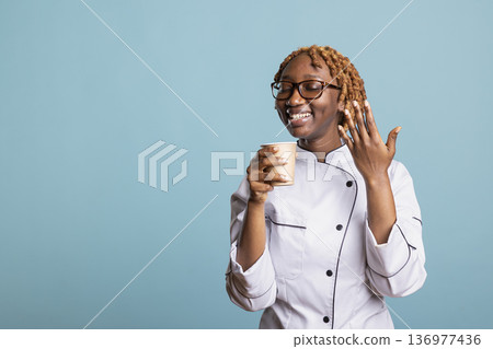 Smiling female chef takes quiet coffee break after demanding day of cooking, holding cup with satisfaction. Young culinary professional enjoys warm drink, feeling comfort and relief after busy shift. Smiling female chef takes quiet coffee break after demanding day of cooking, holding cup with satisfaction. Young culinary professional enjoys warm drink, feeling comfort and relief after busy shift. 136977436
