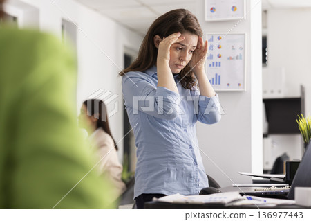Caucasian woman massaging head in exhaustion, reviewing documents on laptop during demanding workday. Tired female employee shows signs of burnout and mental fatigue in modern corporate office. Caucasian woman massaging head in exhaustion, reviewing documents on laptop during demanding workday. Tired female employee shows signs of burnout and mental fatigue in modern corporate office. 136977443