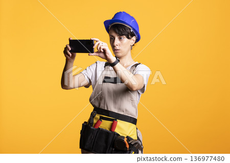 Adult female builder in safety helmet captures photo using smartphone with focused attention. Caucasian builder in construction attire taking picture and visually inspecting renovation site. 136977480