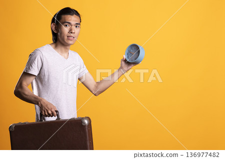Man holds clock and briefcase with worried expression, reacting to fear of missing business meeting. Traveler looking rushed and anxious, balancing time pressure and travel urgency in tense moment. Man holds clock and briefcase with worried expression, reacting to fear of missing business meeting. Traveler looking rushed and anxious, balancing time pressure and travel urgency in tense moment. 136977482