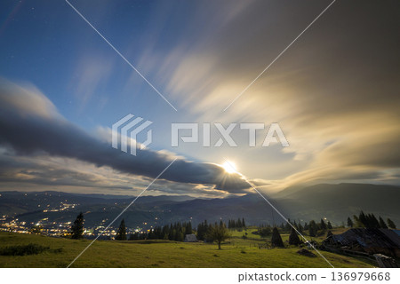 Summer night mountain panorama. Green grassy mountain clearing, spruce trees on blue evening sky copy space background, bright road with moving cars and dwelling lights below in valley. Summer night mountain panorama. Green grassy mountain clearing, spruce trees on blue evening sky copy space background, bright road with moving cars and dwelling lights below in valley. 136979668