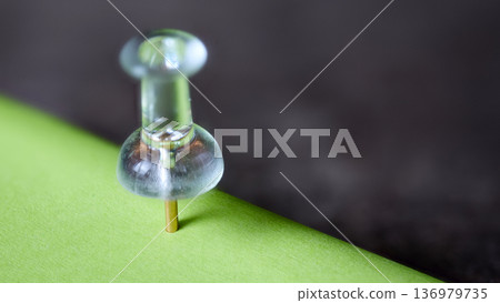 Macro shot of a clear plastic map pin inserted into the edge of a vibrant green sticky note 136979735