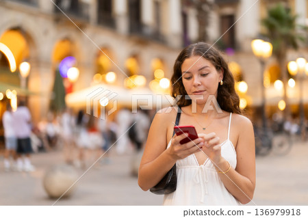 In evening, smiling girl with smartphone walks along city street Placa Real In evening, smiling girl with smartphone walks along city street Placa Real 136979918