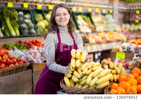 Young female seller holding bananas standing by counter in grocery market Young female seller holding bananas standing by counter in grocery market 136979952