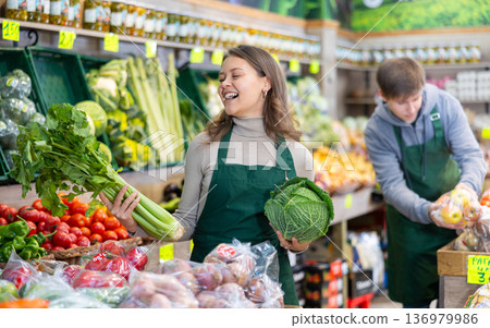 Young female seller holding cabbage and celery standing by counter in grocery market Young female seller holding cabbage and celery standing by counter in grocery market 136979986