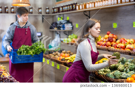 Young female seller holding artichoke standing by counter in grocery market Young female seller holding artichoke standing by counter in grocery market 136980013