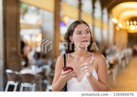 In evening, smiling girl with smartphone walks along city street Placa Real 136980076