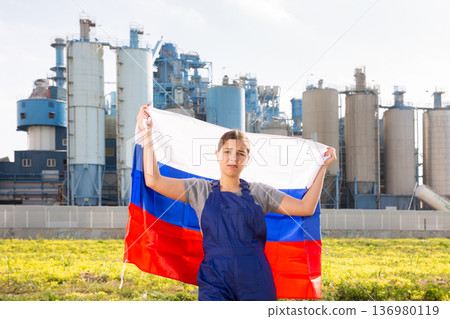 Sad young woman worker with flag of Russia against background of factory Sad young woman worker with flag of Russia against background of factory 136980119