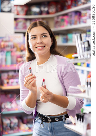 Positive attractive woman holding and comparing two lipsticks while shopping in beauty store Positive attractive woman holding and comparing two lipsticks while shopping in beauty store 136980156