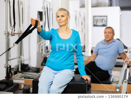 Elderly woman performing toning Pilates exercise for arms during group class in rehabilitation center Elderly woman performing toning Pilates exercise for arms during group class in rehabilitation center 136980174