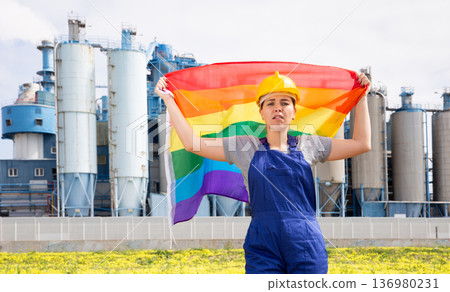 Portrait of upset girl in construction overalls with the LGBT flag on the background of modern factory 136980231