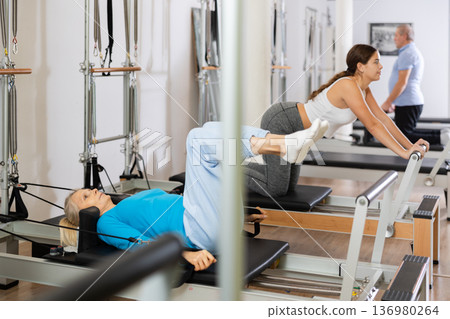 Focused elderly woman lying while doing Pilates tone exercise for back on reformer bed equipment in gym Focused elderly woman lying while doing Pilates tone exercise for back on reformer bed equipment in gym 136980264